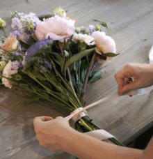 En person binder en bukett med blommor med ett band på ett träbord.